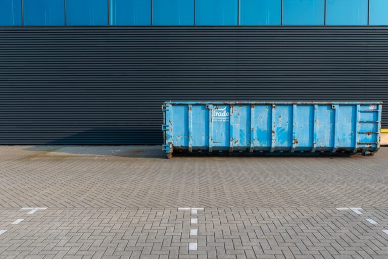 Blue metal dumpster with rust spots in front of a dark blue wall on a brick-paved lot.