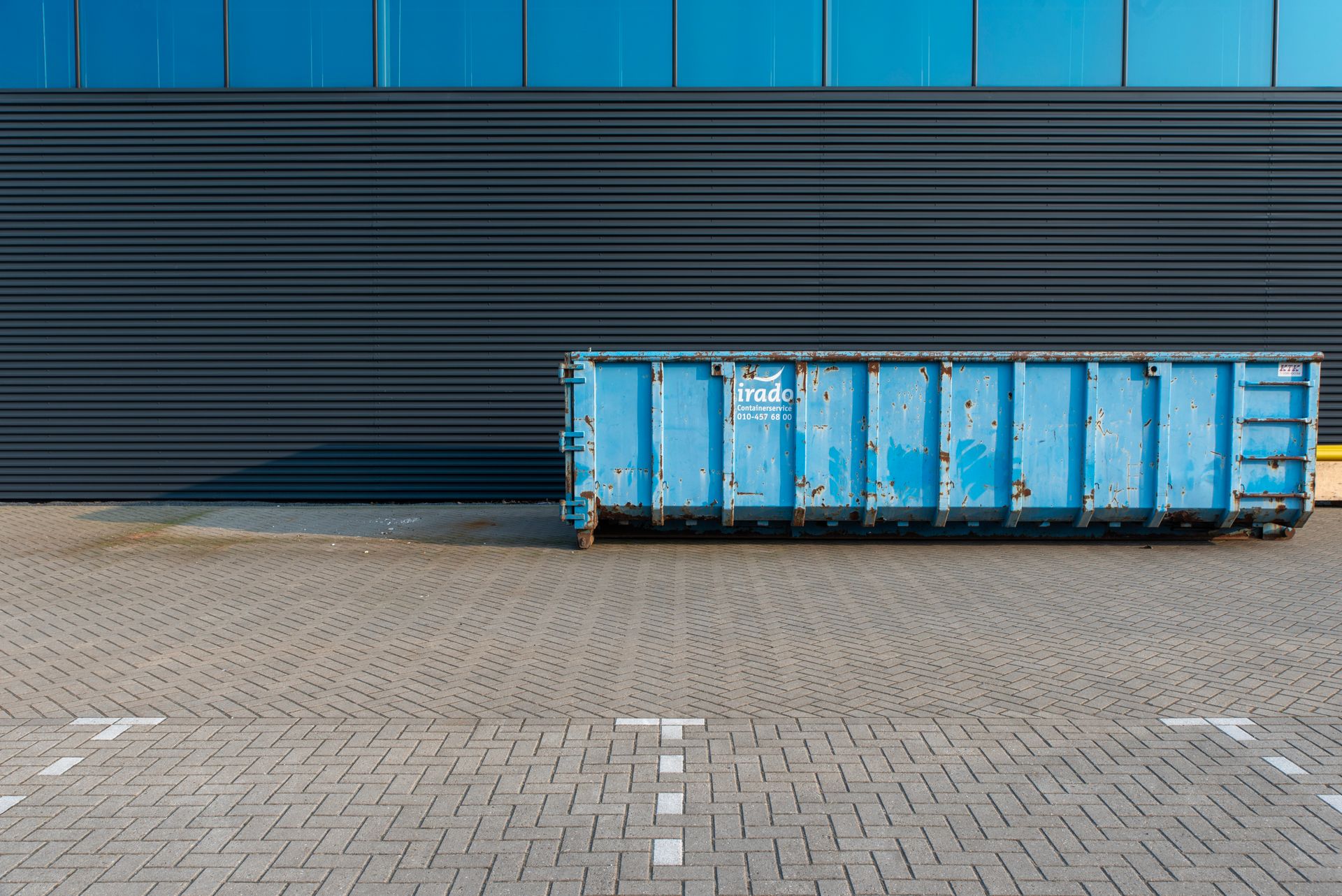 Blue metal dumpster with rust spots in front of a dark blue wall on a brick-paved lot.